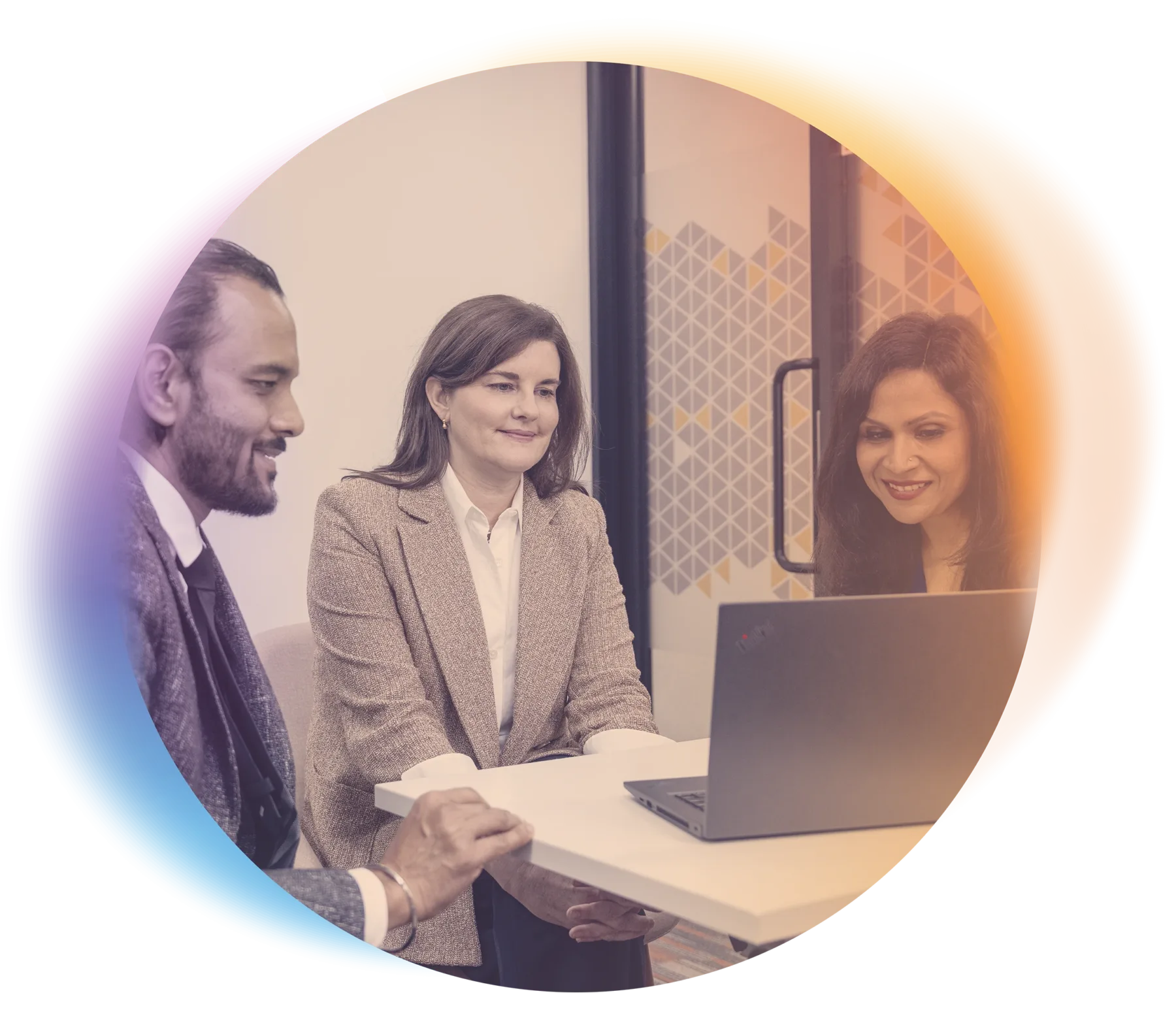 Three professionally dressed people sit together at a small table in an office, looking at a laptop and smiling. The photo is framed within a colorful, gradient oval shape.