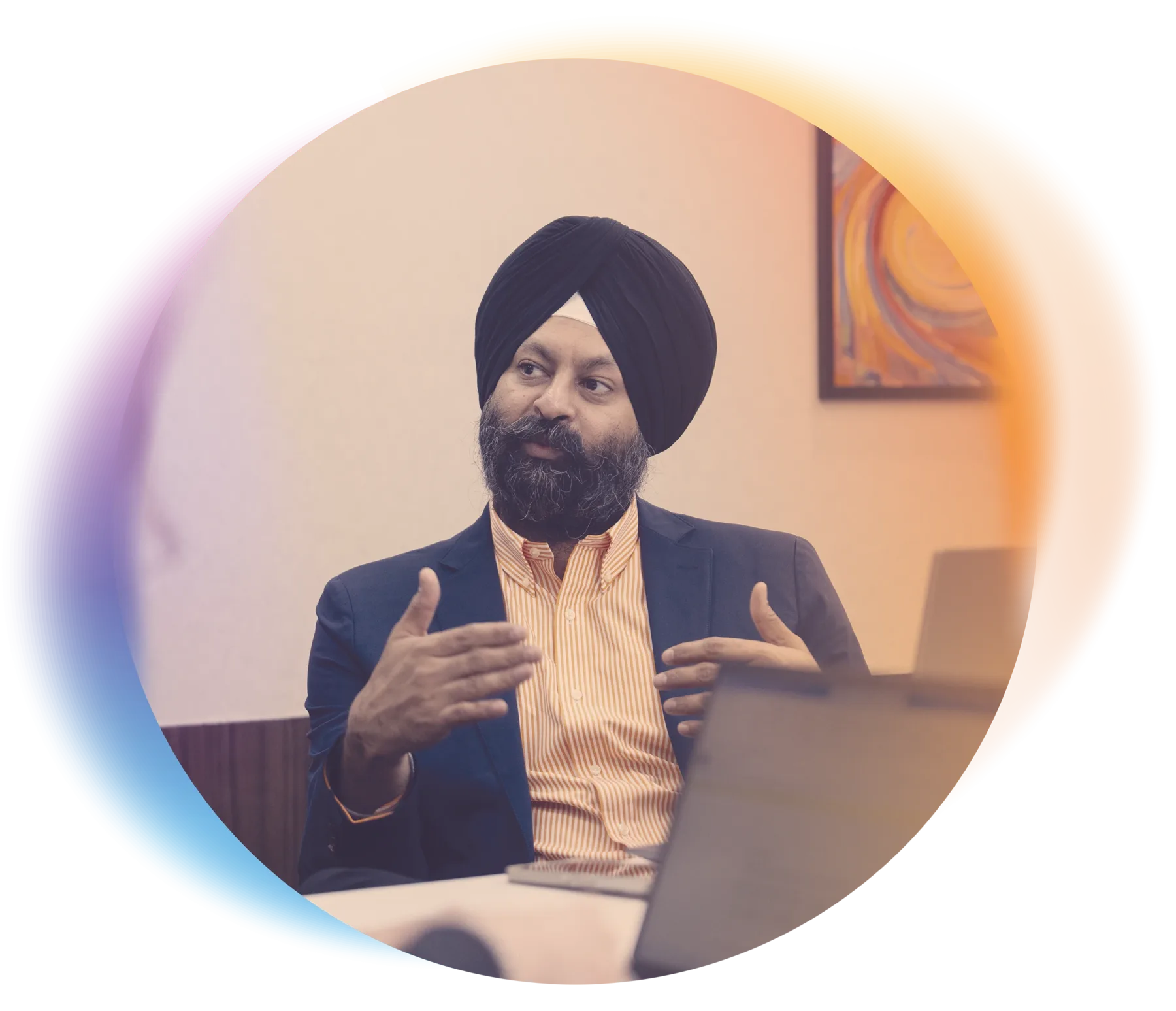 Man wearing a turban and business attire sits at a table in an office, gesturing with his hands while speaking. A colorful gradient shape frames the photo.
